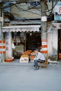 An outdoor market scene with a man sitting on a wooden chair in front of a small fruit stand. The stand displays various fruits, including oranges and mangoes, in baskets and boxes. There are signs in another language surrounding the entrance of the shop. The structure is rustic and the wires above hang loosely.