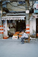 An outdoor market scene with a man sitting on a wooden chair in front of a small fruit stand. The stand displays various fruits, including oranges and mangoes, in baskets and boxes. There are signs in another language surrounding the entrance of the shop. The structure is rustic and the wires above hang loosely.