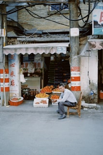 An outdoor market scene with a man sitting on a wooden chair in front of a small fruit stand. The stand displays various fruits, including oranges and mangoes, in baskets and boxes. There are signs in another language surrounding the entrance of the shop. The structure is rustic and the wires above hang loosely.