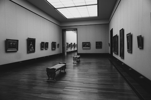 A spacious art gallery with wooden floors and walls adorned with framed paintings. The room is well-lit with a large overhead skylight, and two wooden benches are positioned in the center.