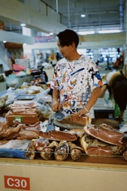 A man wearing a colorful, patterned shirt stands behind a market stall in an indoor setting. The stall is laden with various types of cured meats, including what appears to be prosciutto and other charcuterie items. The stall is marked with a red label reading 'C30.' In the background, there are shelves and other market-goers.