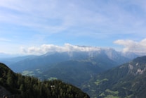 A panoramic view of mountainous landscapes with a clear blue sky above. The peaks are partially covered with clouds, and lush green forests blanket the lower slopes. In the distance, various shades of blue and green create a layered effect, emphasizing the vastness of the scene.