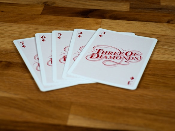 A set of playing cards is fanned out on a wooden surface, showing the 7, 6, 5, 4, and 3 of diamonds, with the 3 card prominently featured.