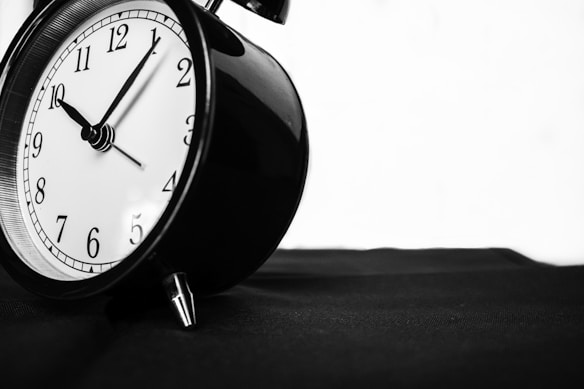 A classic black and white alarm clock with a round face showing the time as 10:10, placed on a dark surface with a minimalist background.