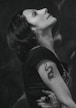 A woman is captured in profile, gazing upward with a serene expression. Her short, dark hair is styled neatly, and she wears a black shirt. A tattoo of a dragon is visible on her upper arm. The lighting accentuates her features, creating a contemplative mood against a softly blurred background.