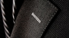 A close-up of a textured suit lapel with detailed stitching, accompanied by a patterned tie in the background.