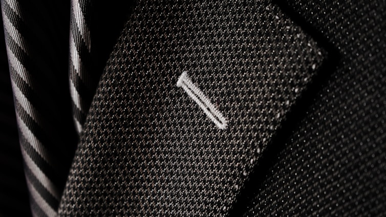 A close-up of a textured suit lapel with detailed stitching, accompanied by a patterned tie in the background.