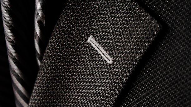 A close-up of a textured suit lapel with detailed stitching, accompanied by a patterned tie in the background.