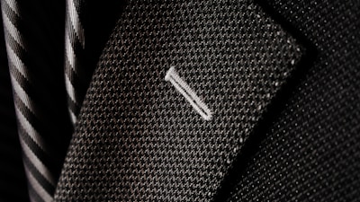 A close-up of a textured suit lapel with detailed stitching, accompanied by a patterned tie in the background.
