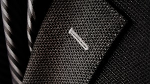 A close-up of a textured suit lapel with detailed stitching, accompanied by a patterned tie in the background.