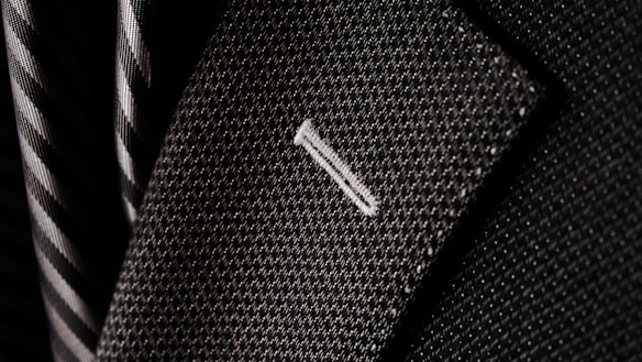 A close-up of a textured suit lapel with detailed stitching, accompanied by a patterned tie in the background.