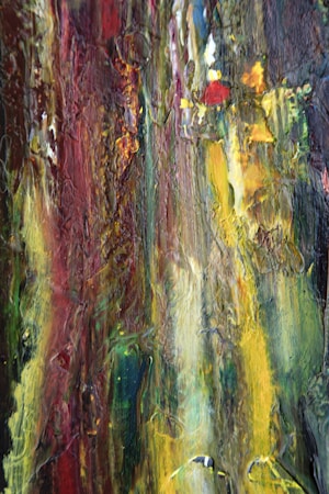 A close-up of a textured abstract painting featuring a mix of vibrant colors including red, yellow, green, and blue. The paint appears thickly applied, creating a rough surface with visible brush strokes and layering. The colors blend together in a vertical orientation, suggesting a sense of movement and energy.