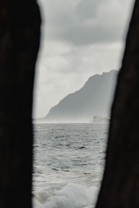 A coastal landscape featuring a large body of water with visible waves and a distant silhouette of a mountain. The image is framed by darker objects in the foreground that partially obscure the view, creating a natural vignette effect. The sky is overcast with soft, diffused lighting suggesting a calm and subdued atmosphere.