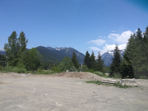 A scenic landscape featuring a dirt lot in the foreground with a pile of dirt and some construction materials. In the middle ground, there are lush green trees and utility poles. In the background, a range of mountains can be seen with some snow atop them, under a clear blue sky with a few clouds.