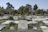A cemetery with numerous tombstones arranged in neat rows, surrounded by green grass and a few scattered trees. In the background, a small building with a tiled roof and glass walls is visible, against a clear blue sky.
