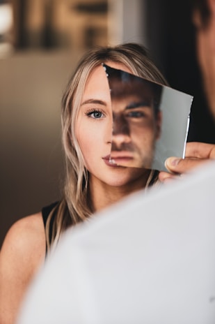 A person holds a small mirror fragment over the face of a woman, creating an illusion where half of her face appears replaced by the reflection of a man's face. Her blonde hair and blue eyes are visible.
