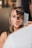 A person holds a small mirror fragment over the face of a woman, creating an illusion where half of her face appears replaced by the reflection of a man's face. Her blonde hair and blue eyes are visible.