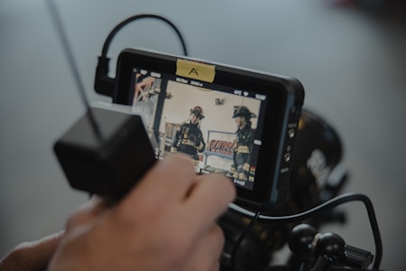 A close-up of a person holding a monitor displaying a video feed. The screen shows two individuals in firefighter uniforms inside a building. The image captures the monitoring equipment setup, including cables and a control box.