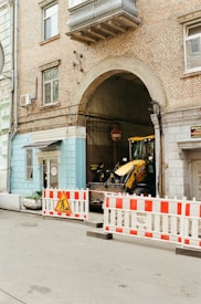 A construction site setup with a backhoe loader parked in an archway entrance of an old brick building. The area is cordoned off with red and white barricades, and a construction sign indicating work in progress. Above the archway, there are windows and an AC unit attached to the wall.