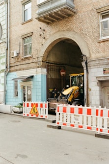 A construction site setup with a backhoe loader parked in an archway entrance of an old brick building. The area is cordoned off with red and white barricades, and a construction sign indicating work in progress. Above the archway, there are windows and an AC unit attached to the wall.