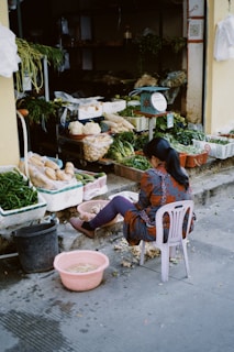 A person is seated on a plastic chair peeling vegetables in front of a small market stall. The stall contains various fresh produce, including green leafy vegetables, cabbages, and potatoes in baskets and containers. The ground is scattered with vegetable peelings, and there's a pink basin with water nearby.
