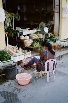 A person is seated on a plastic chair peeling vegetables in front of a small market stall. The stall contains various fresh produce, including green leafy vegetables, cabbages, and potatoes in baskets and containers. The ground is scattered with vegetable peelings, and there's a pink basin with water nearby.