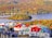 A vibrant village is nestled by a serene lake, surrounded by lush, dense forests in autumn hues of orange, yellow, and green. The buildings in the village feature colorful rooftops in shades of red, blue, and green, creating a picturesque and lively scene against the natural landscape.