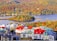 A vibrant village is nestled by a serene lake, surrounded by lush, dense forests in autumn hues of orange, yellow, and green. The buildings in the village feature colorful rooftops in shades of red, blue, and green, creating a picturesque and lively scene against the natural landscape.