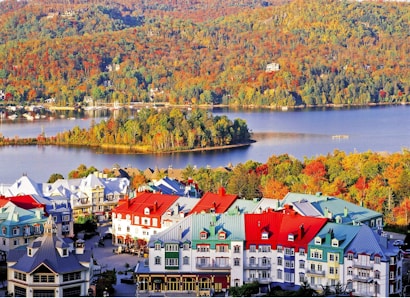 A vibrant village is nestled by a serene lake, surrounded by lush, dense forests in autumn hues of orange, yellow, and green. The buildings in the village feature colorful rooftops in shades of red, blue, and green, creating a picturesque and lively scene against the natural landscape.