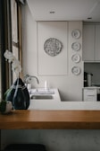 A kitchen interior featuring a modern design with a wooden countertop and a sleek sink. Wall decorations include a larger circular artwork and four smaller plates with artistic designs. An orchid plant and a decorative vase are placed near the window, adding a touch of elegance.