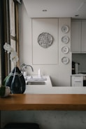 A kitchen interior featuring a modern design with a wooden countertop and a sleek sink. Wall decorations include a larger circular artwork and four smaller plates with artistic designs. An orchid plant and a decorative vase are placed near the window, adding a touch of elegance.
