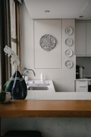 A kitchen interior featuring a modern design with a wooden countertop and a sleek sink. Wall decorations include a larger circular artwork and four smaller plates with artistic designs. An orchid plant and a decorative vase are placed near the window, adding a touch of elegance.