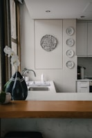 A kitchen interior featuring a modern design with a wooden countertop and a sleek sink. Wall decorations include a larger circular artwork and four smaller plates with artistic designs. An orchid plant and a decorative vase are placed near the window, adding a touch of elegance.