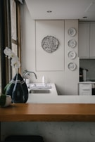 A kitchen interior featuring a modern design with a wooden countertop and a sleek sink. Wall decorations include a larger circular artwork and four smaller plates with artistic designs. An orchid plant and a decorative vase are placed near the window, adding a touch of elegance.