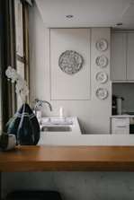 A kitchen interior featuring a modern design with a wooden countertop and a sleek sink. Wall decorations include a larger circular artwork and four smaller plates with artistic designs. An orchid plant and a decorative vase are placed near the window, adding a touch of elegance.