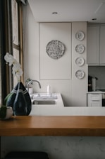 A kitchen interior featuring a modern design with a wooden countertop and a sleek sink. Wall decorations include a larger circular artwork and four smaller plates with artistic designs. An orchid plant and a decorative vase are placed near the window, adding a touch of elegance.