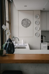 A kitchen interior featuring a modern design with a wooden countertop and a sleek sink. Wall decorations include a larger circular artwork and four smaller plates with artistic designs. An orchid plant and a decorative vase are placed near the window, adding a touch of elegance.