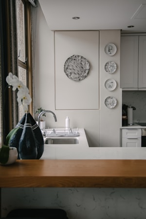 A kitchen interior featuring a modern design with a wooden countertop and a sleek sink. Wall decorations include a larger circular artwork and four smaller plates with artistic designs. An orchid plant and a decorative vase are placed near the window, adding a touch of elegance.