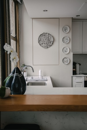 A kitchen interior featuring a modern design with a wooden countertop and a sleek sink. Wall decorations include a larger circular artwork and four smaller plates with artistic designs. An orchid plant and a decorative vase are placed near the window, adding a touch of elegance.