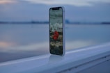A smartphone is placed upright on a railing with a blurry background of a calm body of water and overcast sky. The phone's screen displays a high-resolution image of a red rose on a beach with pebbles.