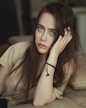 A woman with long hair gazes intently at the camera while resting her head on her hand. Her expression is thoughtful and serene, with natural makeup highlighting her features. She wears a casual light brown top and a black beaded bracelet on her wrist. The background is softly blurred, creating a warm and cozy atmosphere.