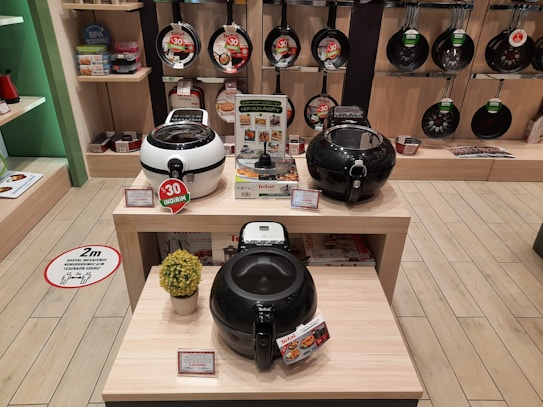 A store display showcasing various kitchen appliances, including several models of air fryers on wooden tables. The background features a wall with hanging frying pans. Discount signs indicate promotions, and a decorative plant is placed next to the appliances.
