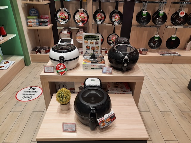 A store display showcasing various kitchen appliances, including several models of air fryers on wooden tables. The background features a wall with hanging frying pans. Discount signs indicate promotions, and a decorative plant is placed next to the appliances.