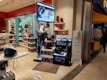 A store display with various home appliances, including vacuum cleaners and irons. The display includes several stacked boxes and products placed on shelves as well as a large digital sign displaying an advertisement. The store is well-lit with a clean, organized layout and there are people in the background near the entrance.