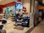 A store display with various home appliances, including vacuum cleaners and irons. The display includes several stacked boxes and products placed on shelves as well as a large digital sign displaying an advertisement. The store is well-lit with a clean, organized layout and there are people in the background near the entrance.