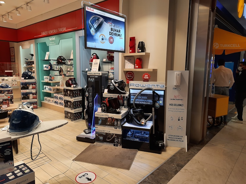 A store display with various home appliances, including vacuum cleaners and irons. The display includes several stacked boxes and products placed on shelves as well as a large digital sign displaying an advertisement. The store is well-lit with a clean, organized layout and there are people in the background near the entrance.