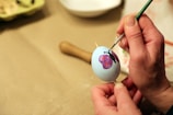 A person is carefully painting a design on a white egg, using a fine brush. The design includes a purple butterfly with red spots. The egg is held steady with a wooden stick in one hand, while the other hand skillfully applies paint.