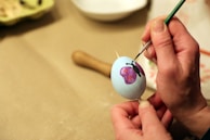 A person is carefully painting a design on a white egg, using a fine brush. The design includes a purple butterfly with red spots. The egg is held steady with a wooden stick in one hand, while the other hand skillfully applies paint.
