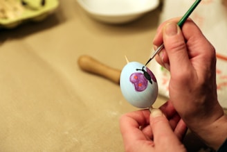 A person is carefully painting a design on a white egg, using a fine brush. The design includes a purple butterfly with red spots. The egg is held steady with a wooden stick in one hand, while the other hand skillfully applies paint.