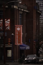 A street scene in Chinatown featuring a brick building with a vertical sign with the word 'Chinatown' in both English and Chinese characters. Red lanterns hang from a pole next to the building. A person is walking near a parked car under dim lighting, suggesting an overcast day.
